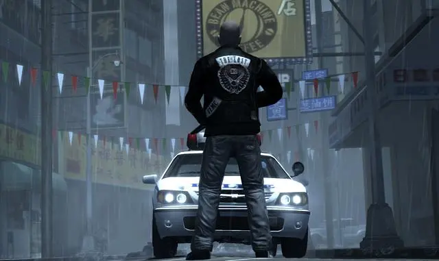 Un membre du club de motards The Lost vu de dos, blouson de cuir avec l'écusson du gang, debout sous la pluie devant une voiture de police aux phares allumés dans une rue de Chinatown à Liberty City