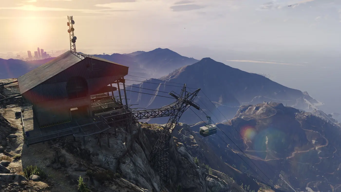 Station du téléphérique au sommet du mont Chiliad dans GTA 5 au coucher du soleil, avec une cabine sur son câble et la skyline de Los Santos dans la brume au loin