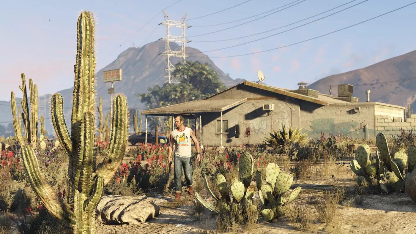 Trevor, personnage de GTA 5 en marcel, marche parmi les cactus et les fleurs sauvages devant une bâtisse isolée du désert de Blaine County au coucher du soleil