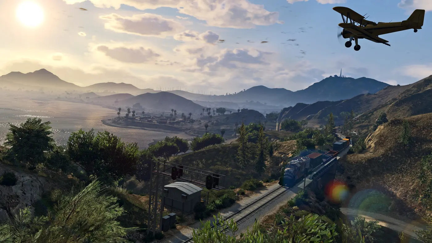 Un petit biplan jaune survole les collines et la côte de Blaine County au lever du soleil dans GTA Online, un train de marchandises passant en contrebas