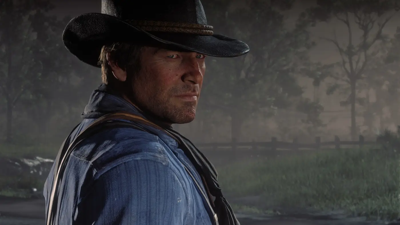 Gros plan d'Arthur Morgan dans Red Dead Redemption 2, visage détaillé, barbe naissante et chapeau usé sous une lumière douce de forêt