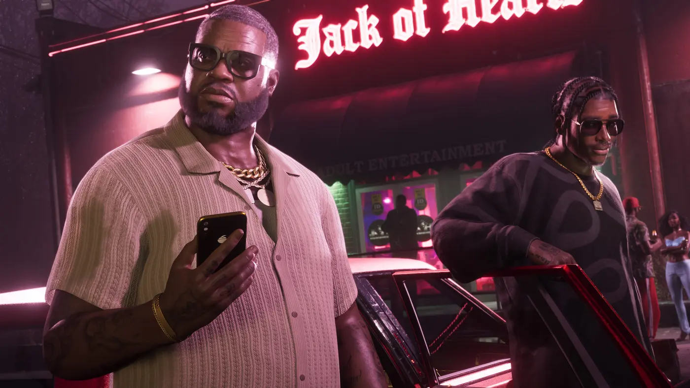 Scène nocturne de GTA VI : un homme corpulent en chemise claire et chaîne en or tient un smartphone à la main devant l'entrée d'un club au néon rouge « Jack of Hearts » portant la mention Adult Entertainment, un second personnage en lunettes noires s'appuie sur la portière d'une voiture sombre